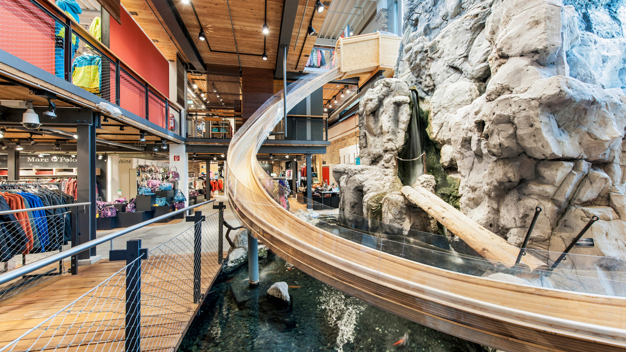Das Bild zeigt das Innere eines Ladens mit einer großen Spiralrutsche, die über künstliches Felsgelände und ein Wasserspiel nach unten führt. Verschiedene Kleidungsstücke sind ausgestellt, und die Decke ist mit Holzakzenten und Industriebeleuchtung versehen.