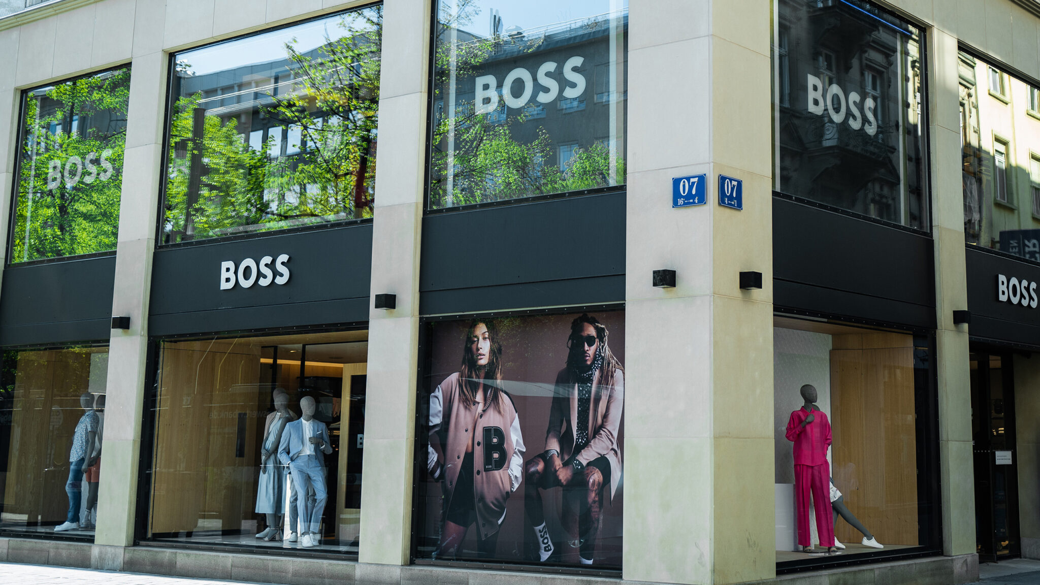 Ein Schaufenster mit großen „BOSS“-Schildern zeigt Schaufensterpuppen in Outfits. In den Fenstern ist eine Werbung mit zwei Personen in modischer Kleidung zu sehen. Das Gebäude steht an einer Straßenecke mit einem blauen Straßenschild.