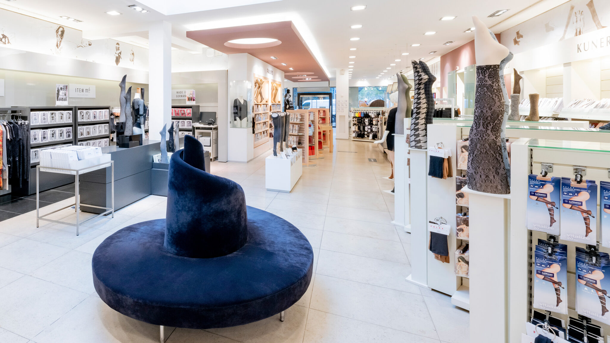 Ein modernes, gut beleuchtetes Ladenlokal mit einem marineblauen, runden Sofa in der Mitte. Auf der rechten Seite präsentieren Schaufensterpuppenbeine Strumpfwaren, und in den Regalen sind verschiedene Produkte ausgestellt. An der Rückwand ist ein großes Frauengesicht abgebildet.