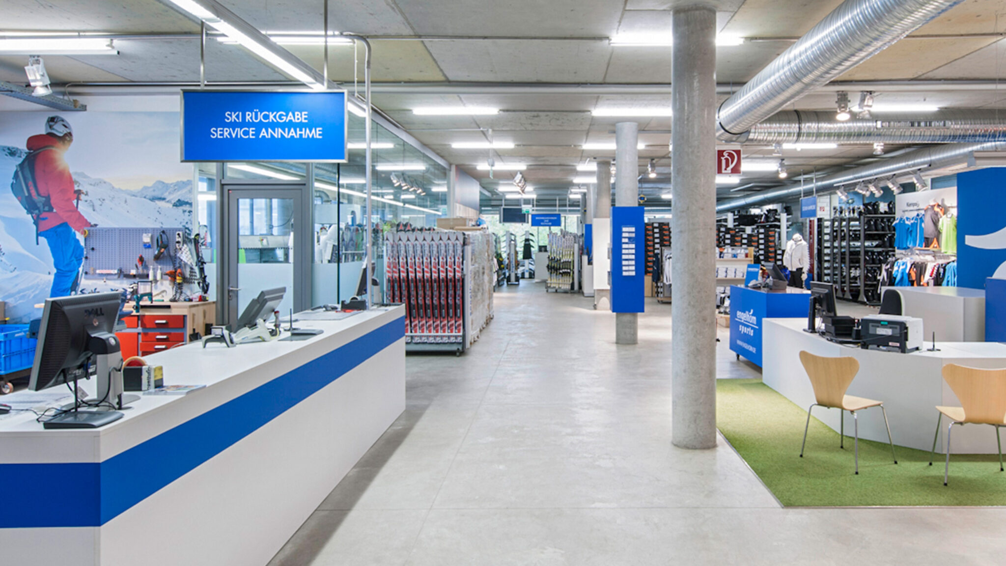 Ein geräumiger Ski- und Outdoor-Ausrüstungsladen mit blauer Beschilderung. Der Innenraum verfügt über Servicetheken, Regale mit Waren und einen Sitzbereich mit Holzstühlen auf einer grünen Matte. Große Bilder von Skifahrern schmücken die Wände.