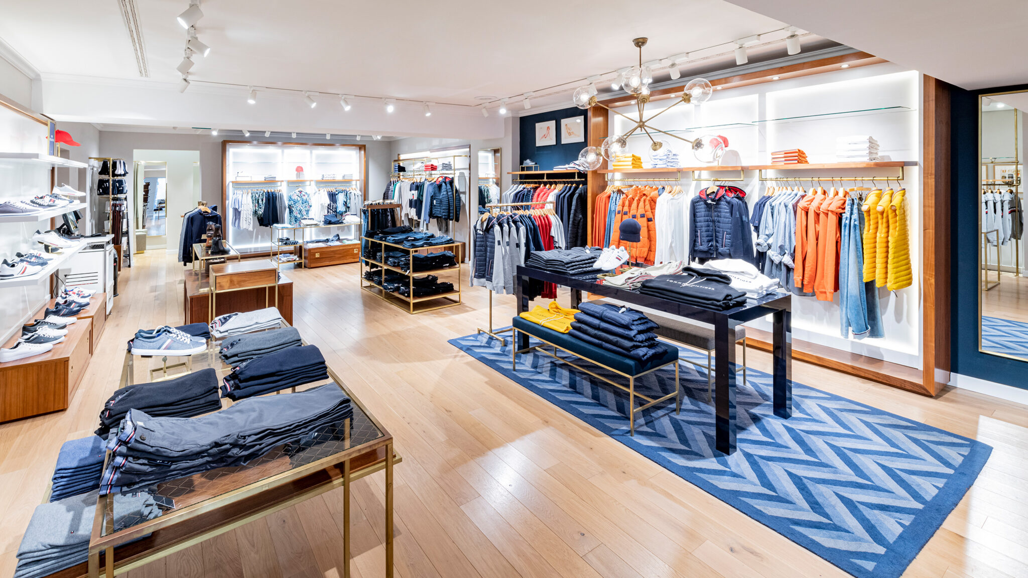 Ein hell erleuchtetes Bekleidungsgeschäft mit Holzboden präsentiert übersichtlich Herrenbekleidung auf Ständern und Tischen. Regale an den Wänden bieten gefaltete Hemden und Jacken in verschiedenen Stilen und Farben. Ein blauer geometrischer Teppich verleiht dem Laden einen modernen Touch.