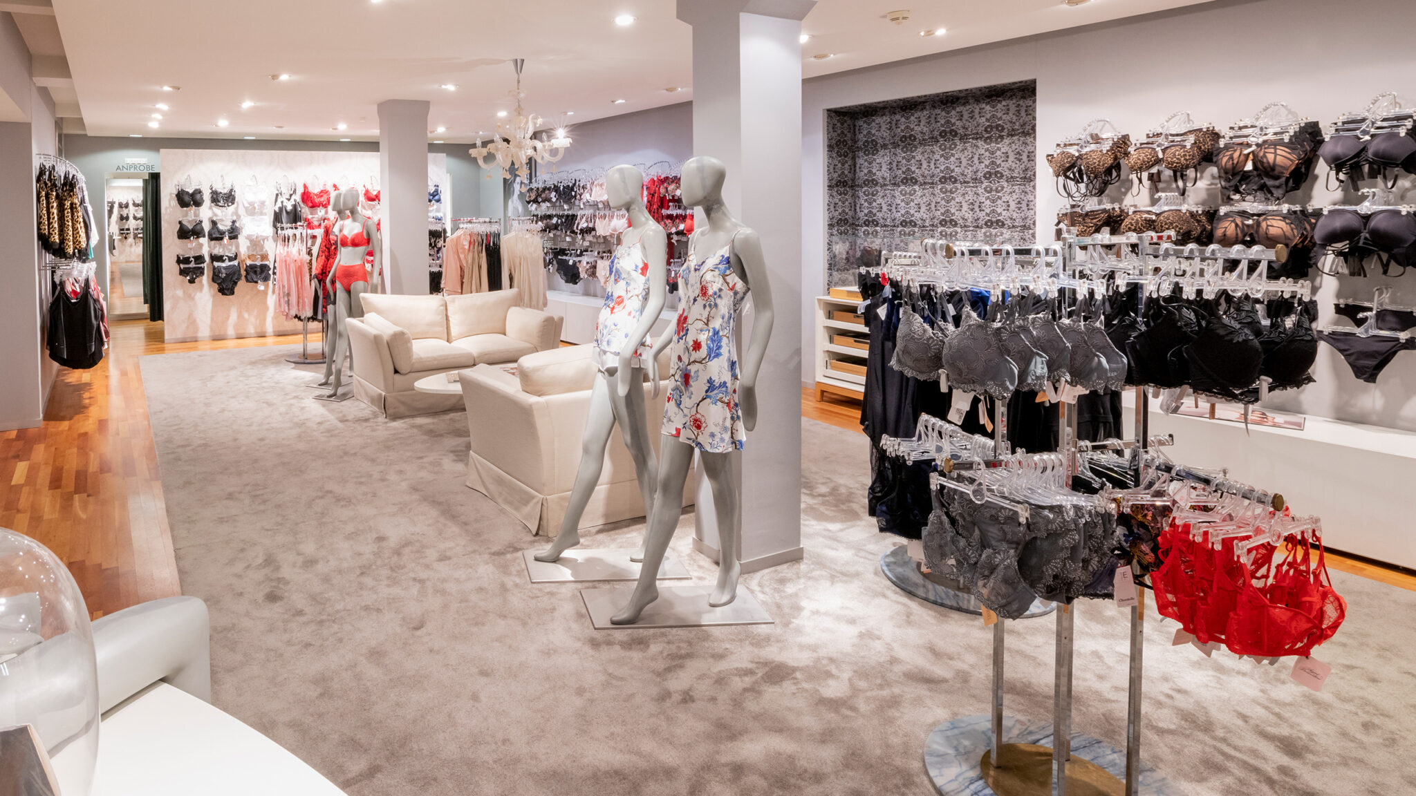 Ein Dessousgeschäft mit grauem Teppichboden und Holzboden. Schaufensterpuppen präsentieren geblümte Nachtwäsche, während an den Regalen verschiedene BHs und Unterwäsche in unterschiedlichen Farben angeboten werden. Zwei weiße Polsterstühle stehen in der Mitte, und an den Wänden befinden sich weitere Auslagen.