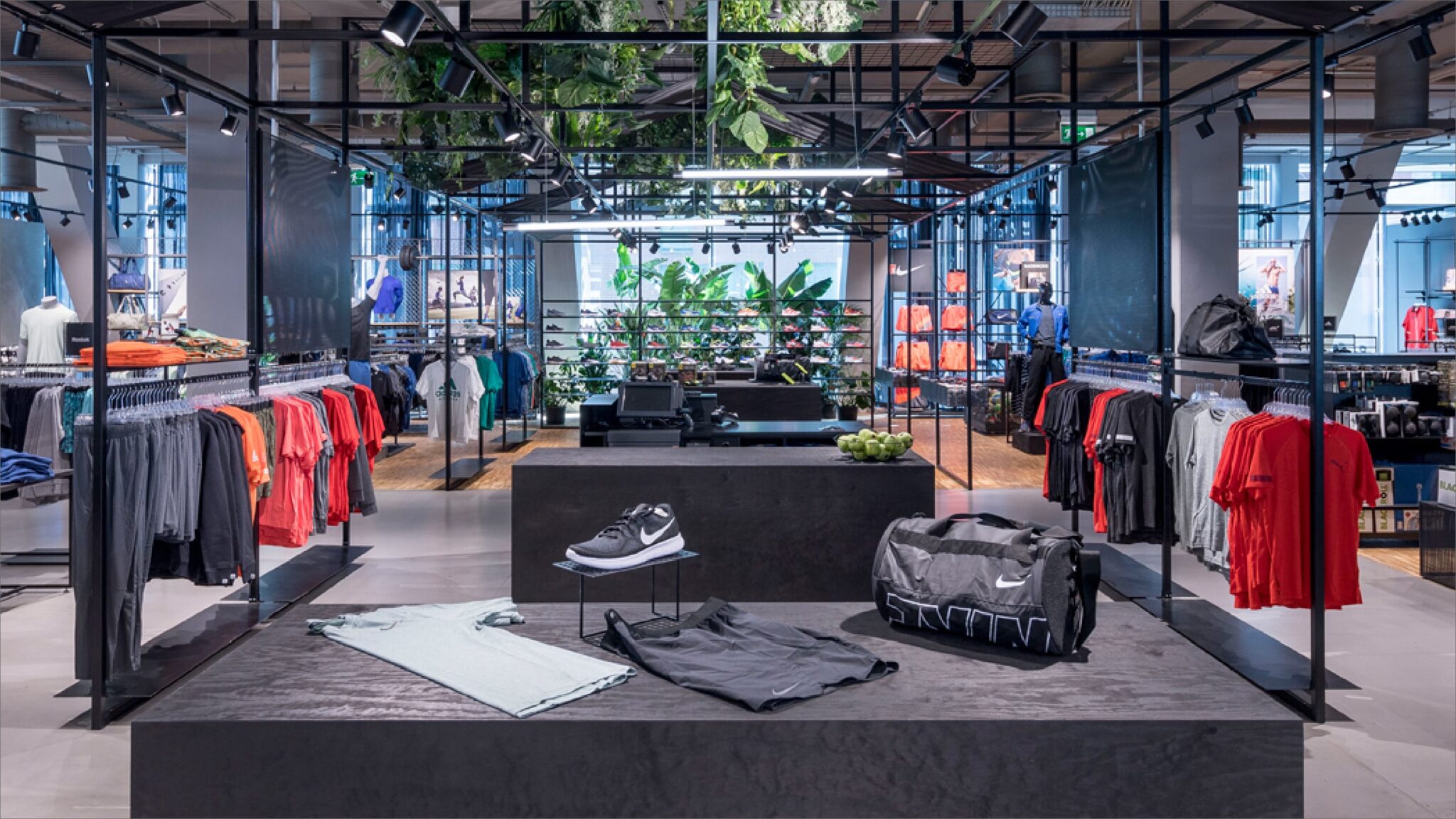 Ein modernes Interieur in einem Sportbekleidungsgeschäft mit Regalen voller farbenfroher Sportbekleidung. Im Vordergrund sind ein graues Hemd, schwarze Shorts, eine Reisetasche und schwarze Turnschuhe zu sehen. Darüber hängen leuchtende Pflanzen von der Decke und werten das Dekor auf.