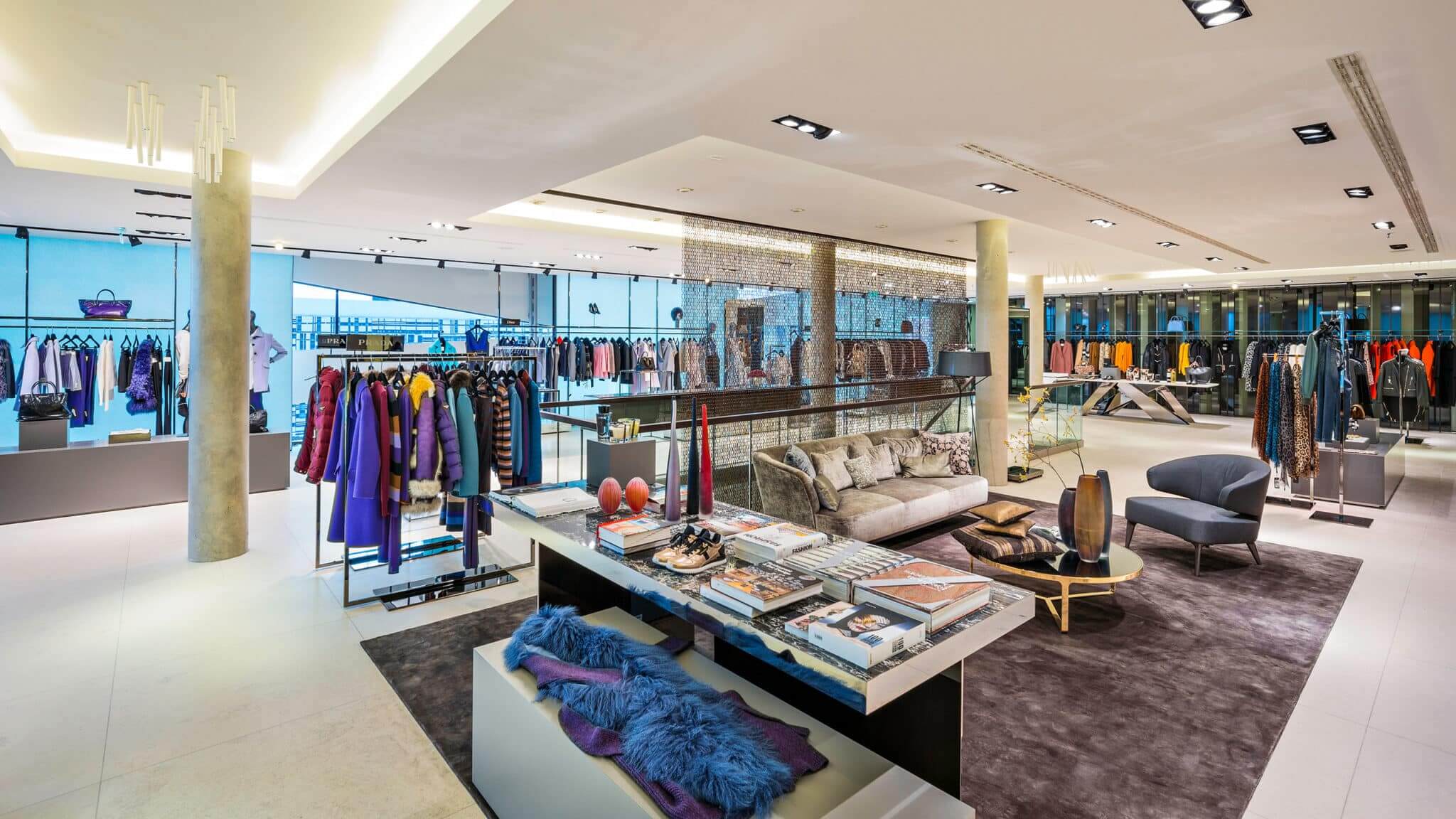 Ein geräumiges, modernes Bekleidungsgeschäft mit farbenfrohen Kleiderständern. Der Innenraum ist mit zeitgenössischen Möbeln, darunter Stühlen und einem Tisch mit Modemagazinen, ausgestattet. Helle Beleuchtung und ein elegantes Design schaffen eine einladende Einkaufsatmosphäre.