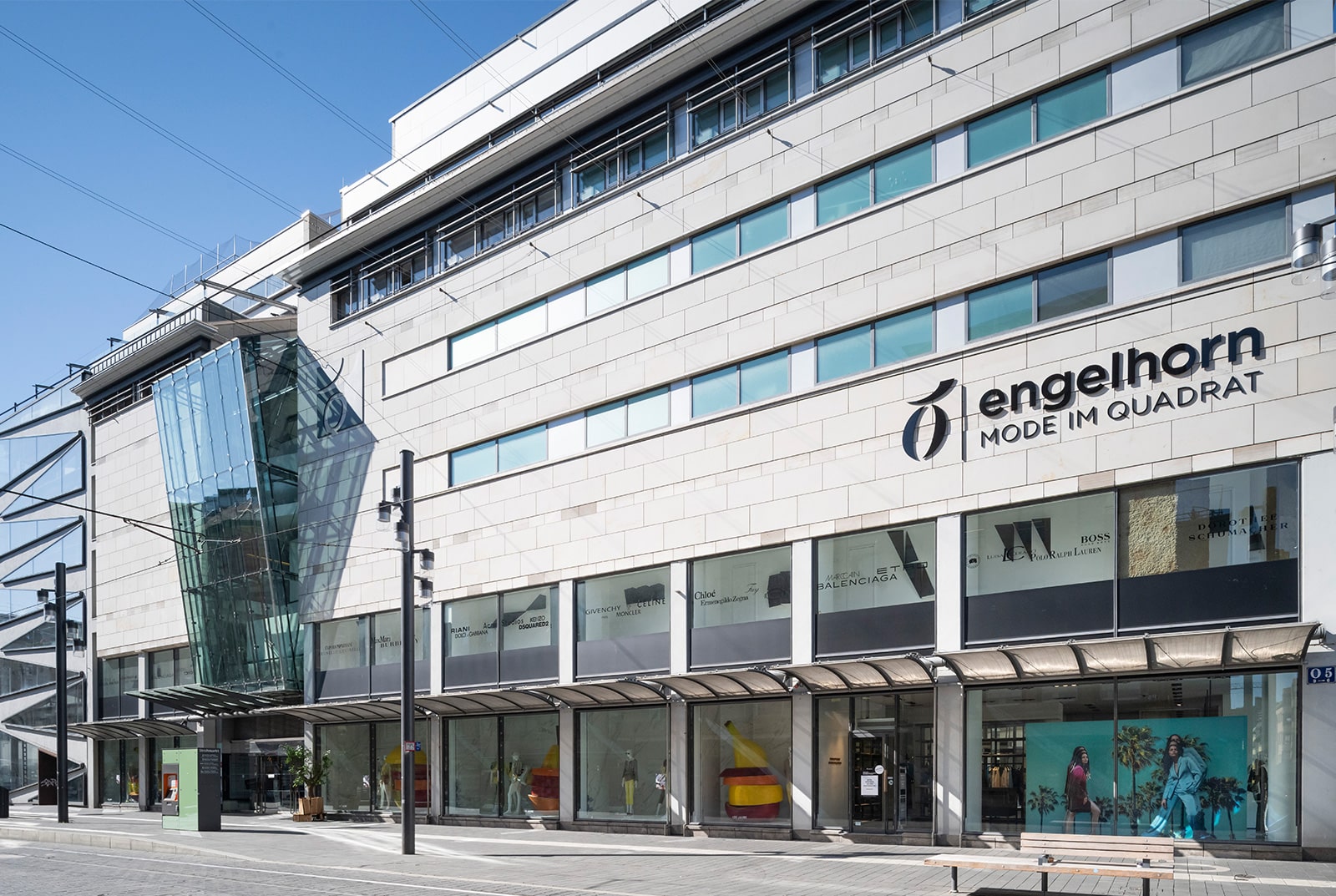 Ein modernes, mehrstöckiges Gebäude mit großen Glasfenstern und einer eckigen Glasfassade. Auf dem Schild steht „engelhorn MODE IM QUADRAT“. Die Straße ist leer und der Himmel klar – ein helles und einladendes Ambiente.