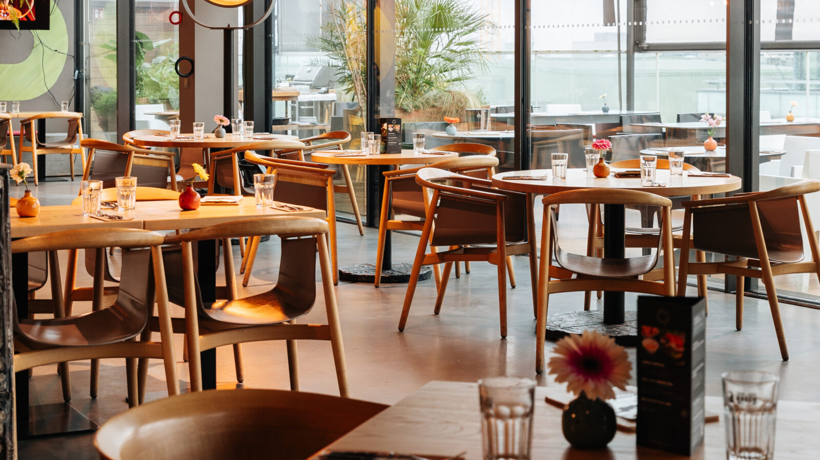 Großzügiges, modernes Restaurant mit Holztischen und -stühlen. Jeder Tisch ist mit Gläsern und kleinen Blumenvasen gedeckt. Große Fenster lassen Tageslicht herein und schaffen eine einladende Atmosphäre.