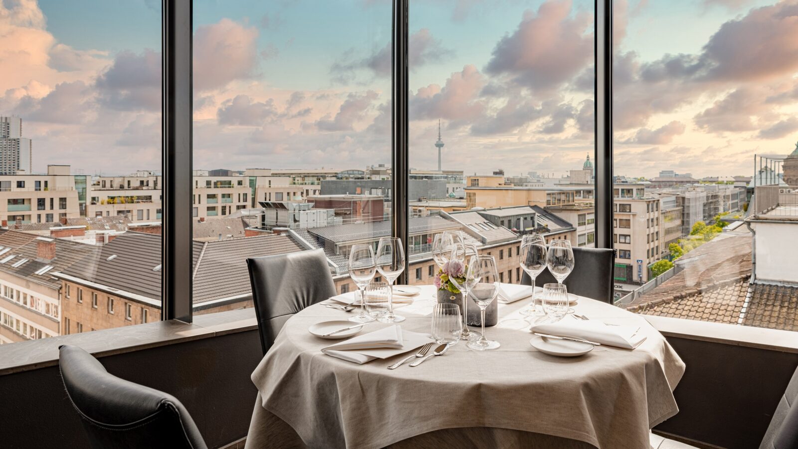 Ein Esstisch für vier Personen mit Weingläsern und Besteck. Durch große Fenster bietet sich ein Panoramablick auf die Stadt. Der Himmel ist von sanften Wolken bedeckt, die von einem warmen Sonnenuntergang erhellt werden und eine ruhige Atmosphäre schaffen.