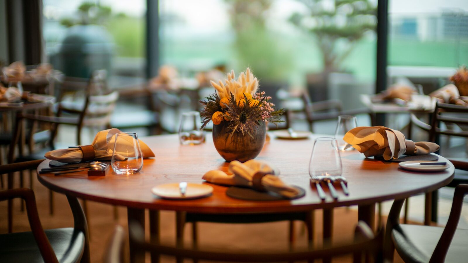 Ein hölzerner Esstisch in einem Restaurant ist elegant mit gefalteten Servietten, Tellern und Gläsern gedeckt. Eine Vase mit Trockenblumen schmückt die Mitte. Im Hintergrund schimmert verschwommenes Grün, und große Fenster lassen natürliches Licht herein.