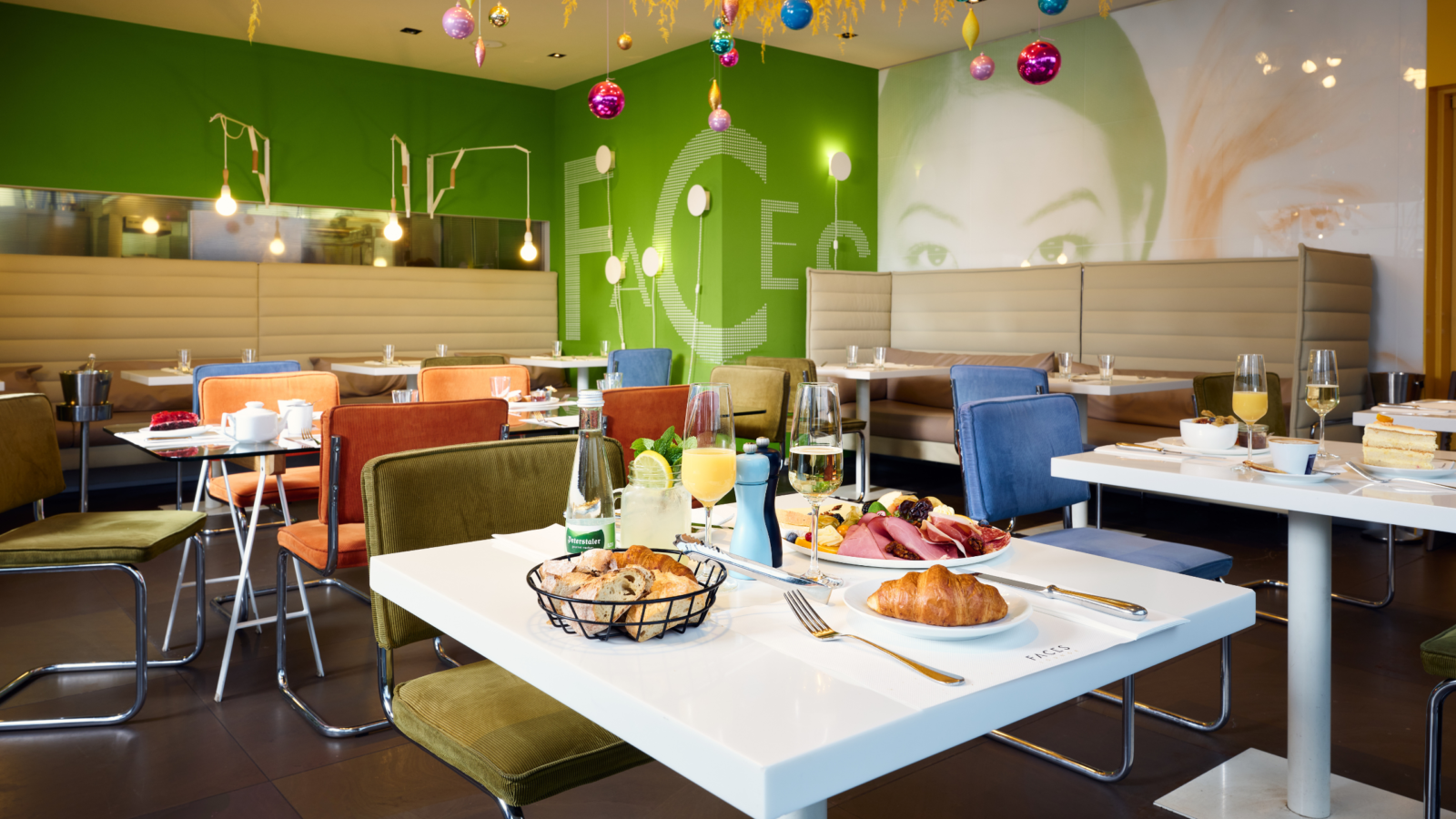 Ein modernes Restaurant mit grünen Wänden, bunten Stühlen und hängenden Ornamenten. Die Tische sind mit Tellern voller Croissants, Fleisch, Obst und Getränken gedeckt. Große Sitzecken stehen an der Wand, und abstrakte Kunst schmückt den Raum.