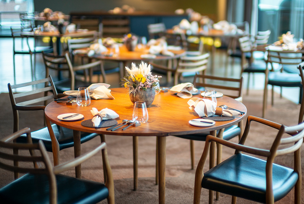 Ein gemütliches Restaurant mit einem runden Holztisch für vier Personen und einem Blumenschmuck. Ordentlich gefaltete Servietten, Gläser und Besteck stehen auf dem Tisch. Holzstühle mit schwarzen Kissen umgeben den Tisch. Weitere Tische sind sichtbar.